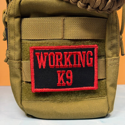 A close-up of a tactical pouch in olive drab green nylon with a black rectangular patch featuring red embroidered text that reads 'WORKING K9'.