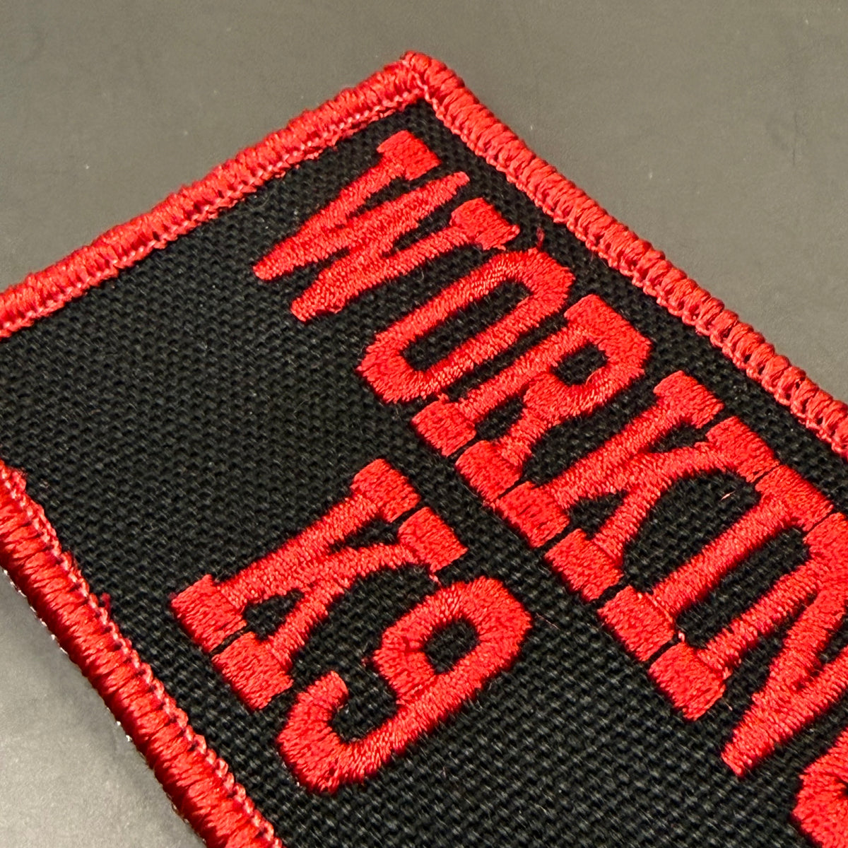 A close-up of a rectangular embroidered patch with a black background, red border, and red text reading 'WORKING 19' on a gray surface.