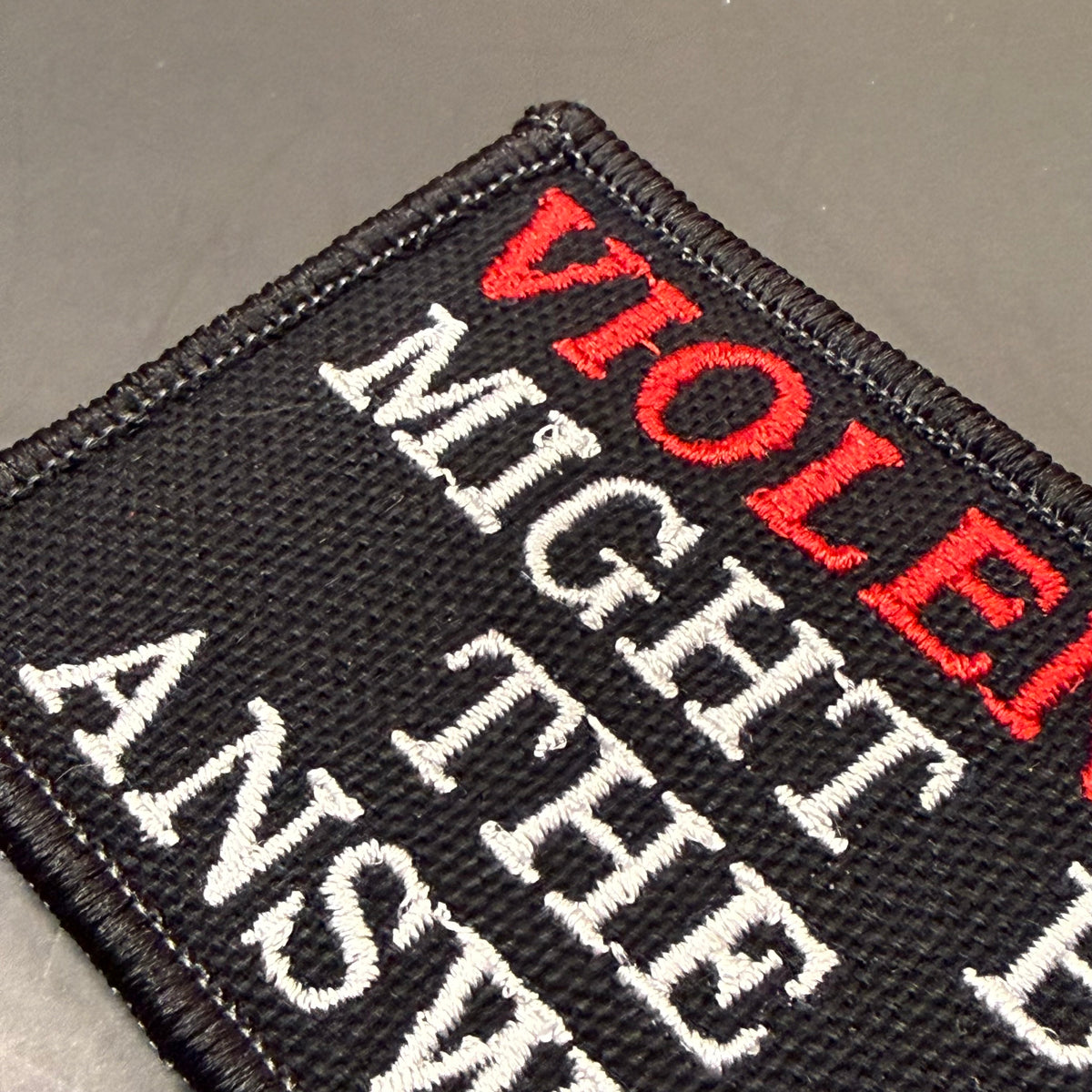 A close-up of a black embroidered patch with white and red text reading 'VIOLENT MIGHT THE ANSWER'.