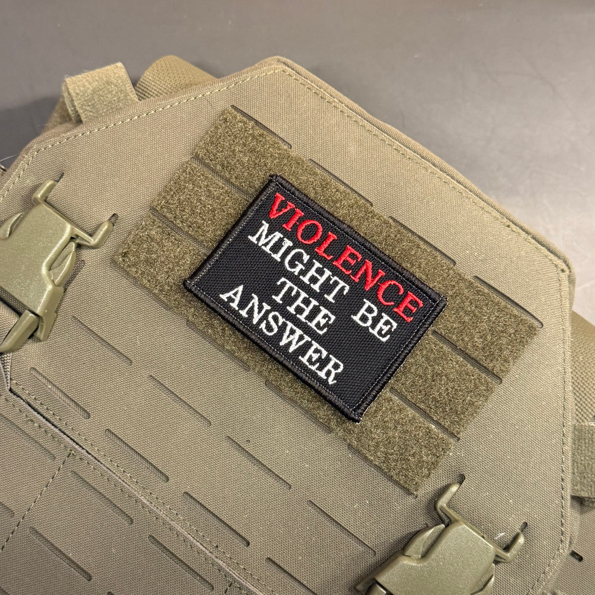 A rectangular embroidered patch with the text 'VIOLENCE MIGHT BE THE ANSWER' in white and red lettering on a black background, attached to tan tactical webbing on a tan bag.