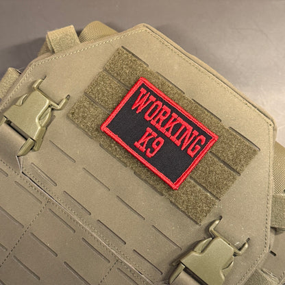 A close-up of a tactical vest or plate carrier in coyote tan, featuring a rectangular black patch with red border and red embroidered text reading 'WORKING K9' on a hook-and-loop section.