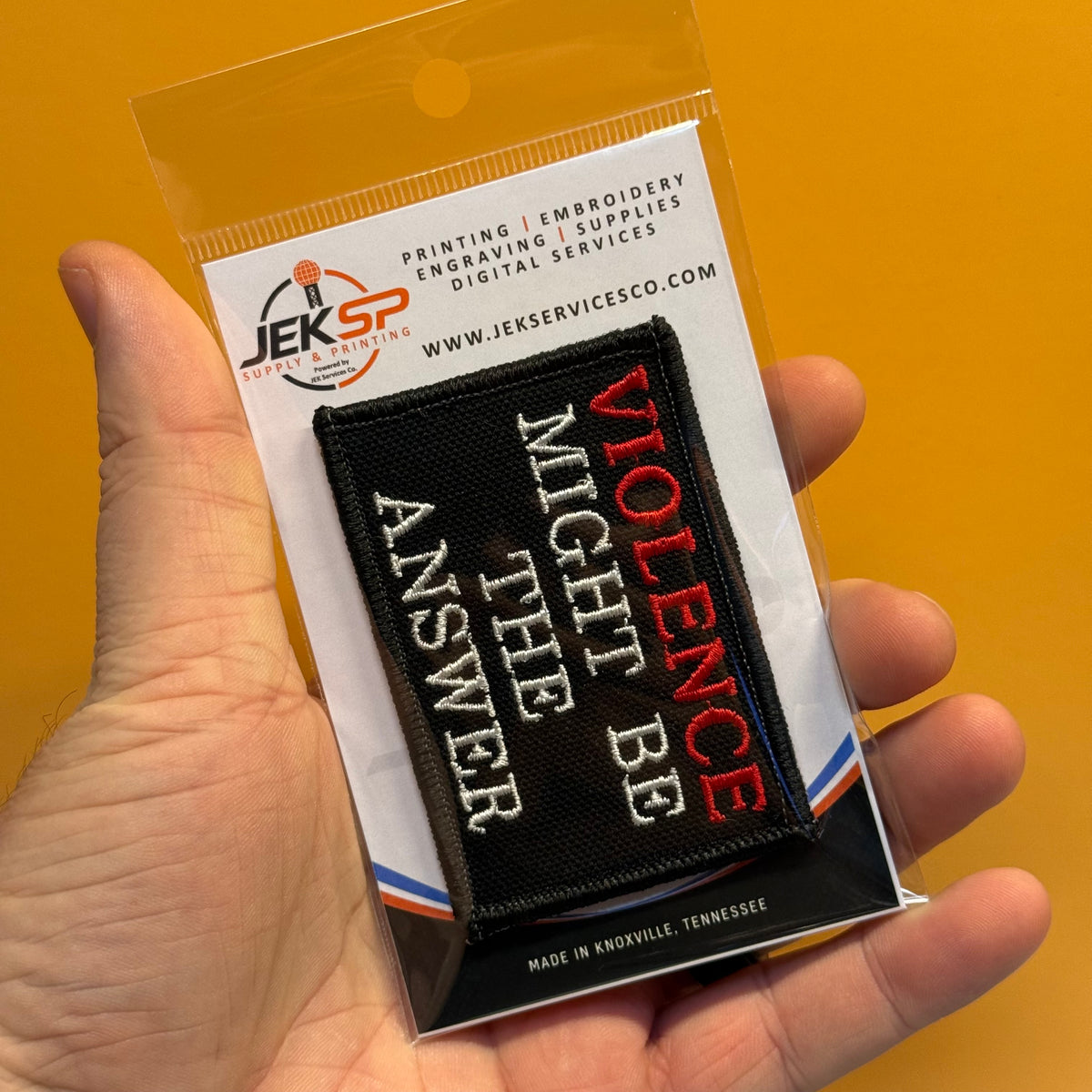 A hand holds a packaged embroidered patch against a solid orange-yellow background. The patch is black with white and red text reading 'VIOLENCE MIGHT BE THE ANSWER'.