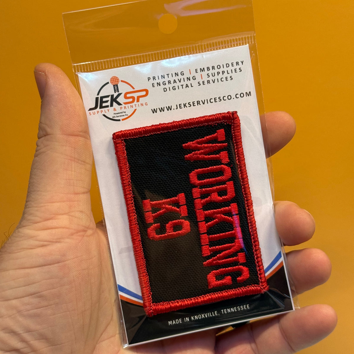 A hand holds a packaged embroidered patch that reads 'WORKING K9' in red on a black background, with a white backing card showing the Jeksp brand and website.