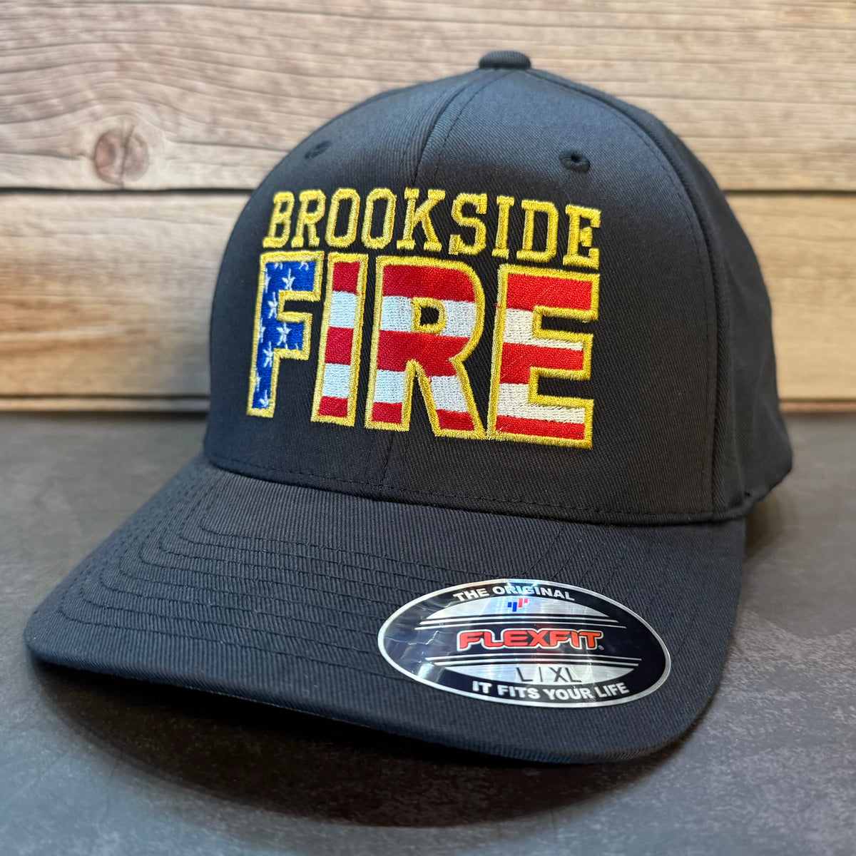 A dark charcoal gray snapback cap with 'Custom FIRE' embroidered on the front. The word 'FIRE' is filled with an American flag pattern, and the cap sits on a dark surface against a rustic wooden background.