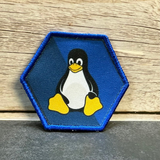 A hexagonal embroidered patch featuring the Tuxedo penguin logo on a blue background with a blue stitched border, placed on a dark surface against a light wood wall.