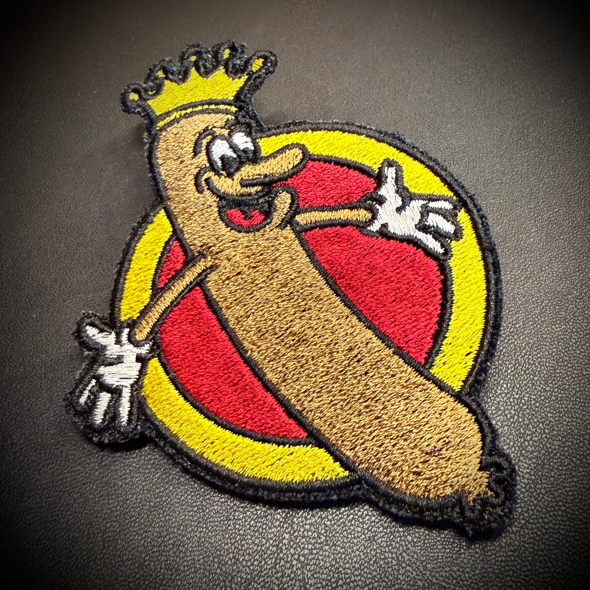 An embroidered patch featuring a cartoon hot dog wearing a gold crown, set against a red and yellow circular background, placed on a dark textured surface.