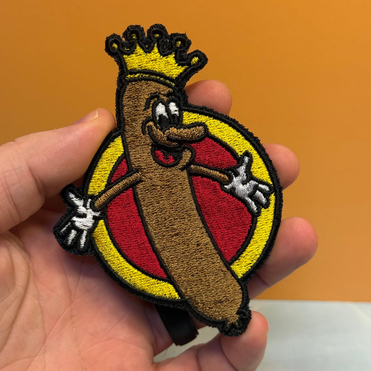 An embroidered patch of a cartoon hot dog character wearing a gold crown, held in a person's hand against an orange background.