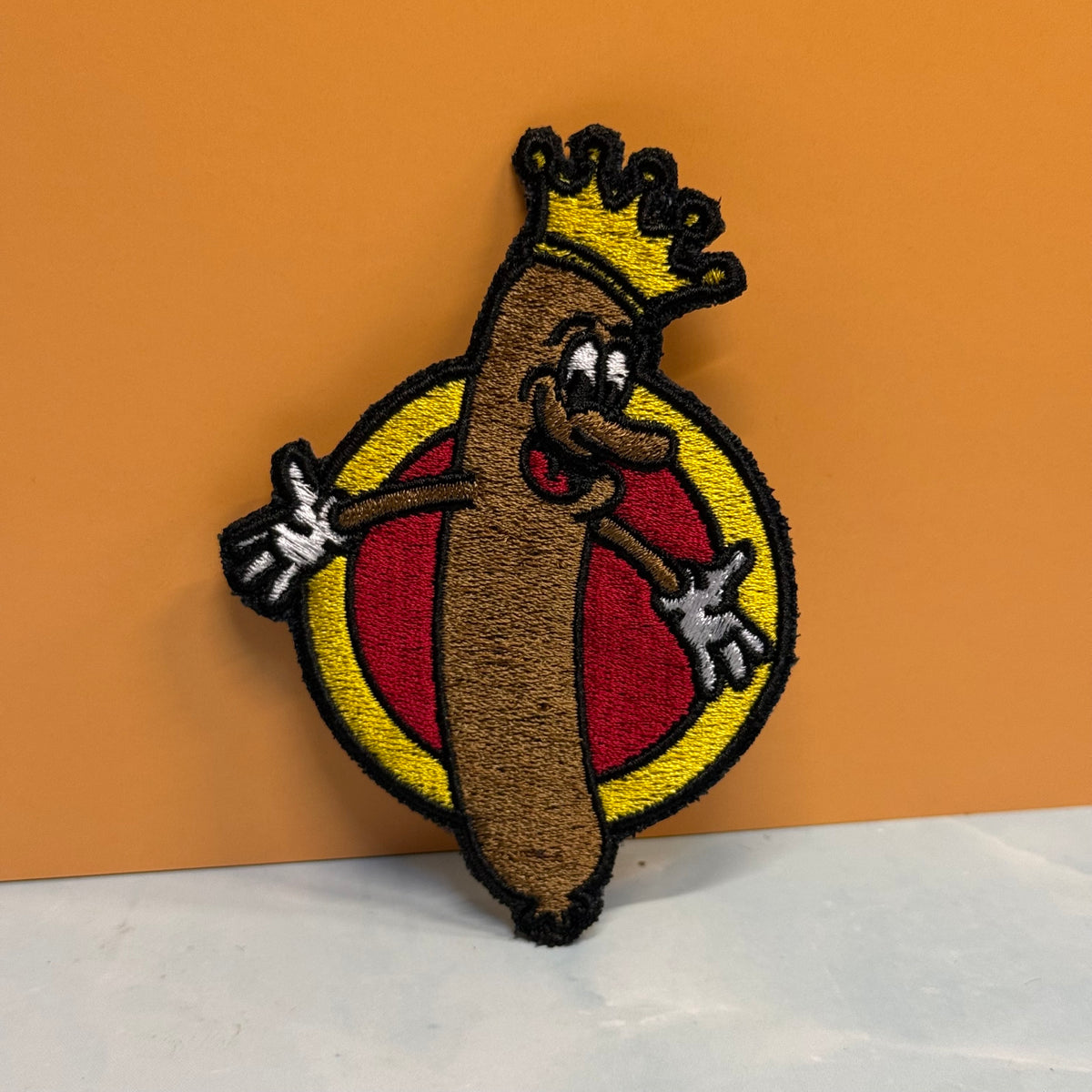 An embroidered patch featuring a cartoon hot dog character wearing a gold crown, set against a yellow and red circular background, displayed on a white surface with an orange backdrop.