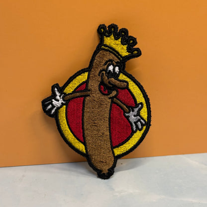 An embroidered patch featuring a cartoon hot dog character wearing a gold crown, set against a yellow and red circular background, displayed on a white surface with an orange backdrop.