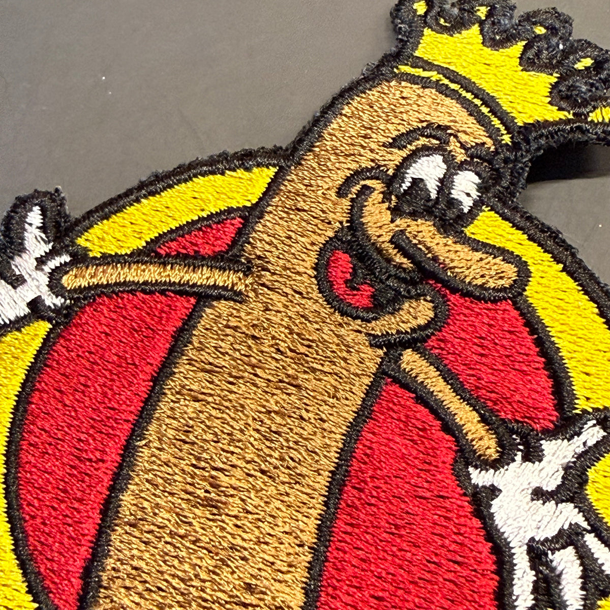 A close-up of a raised tufted rug or patch featuring a cartoon hot dog character wearing a yellow crown and red-and-yellow striped robe, with black outlines and white gloves, photographed on a dark gray background.