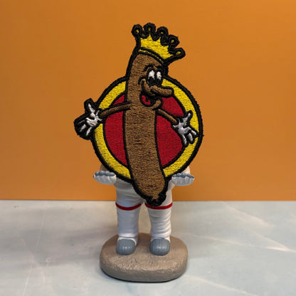 An embroidered patch of a cartoon hot dog character wearing a gold crown, standing in front of a red and yellow circular background, mounted on a small gray base.