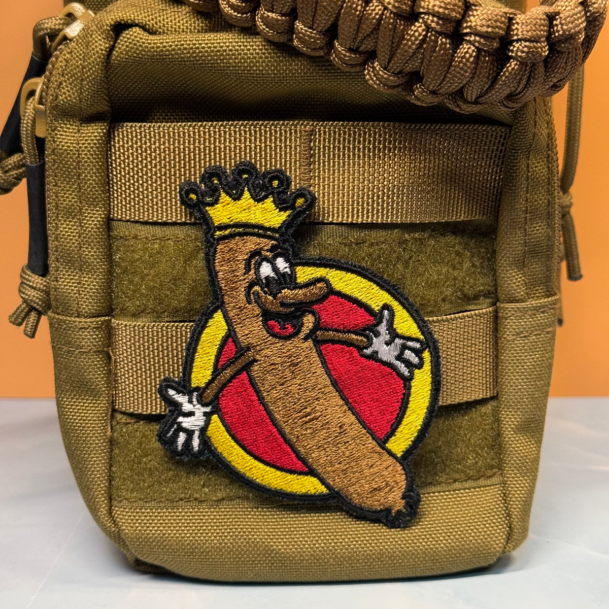 A close-up of a tan tactical pouch featuring a large embroidered patch of a cartoon hot dog wearing a gold crown on a red and yellow circular background.
