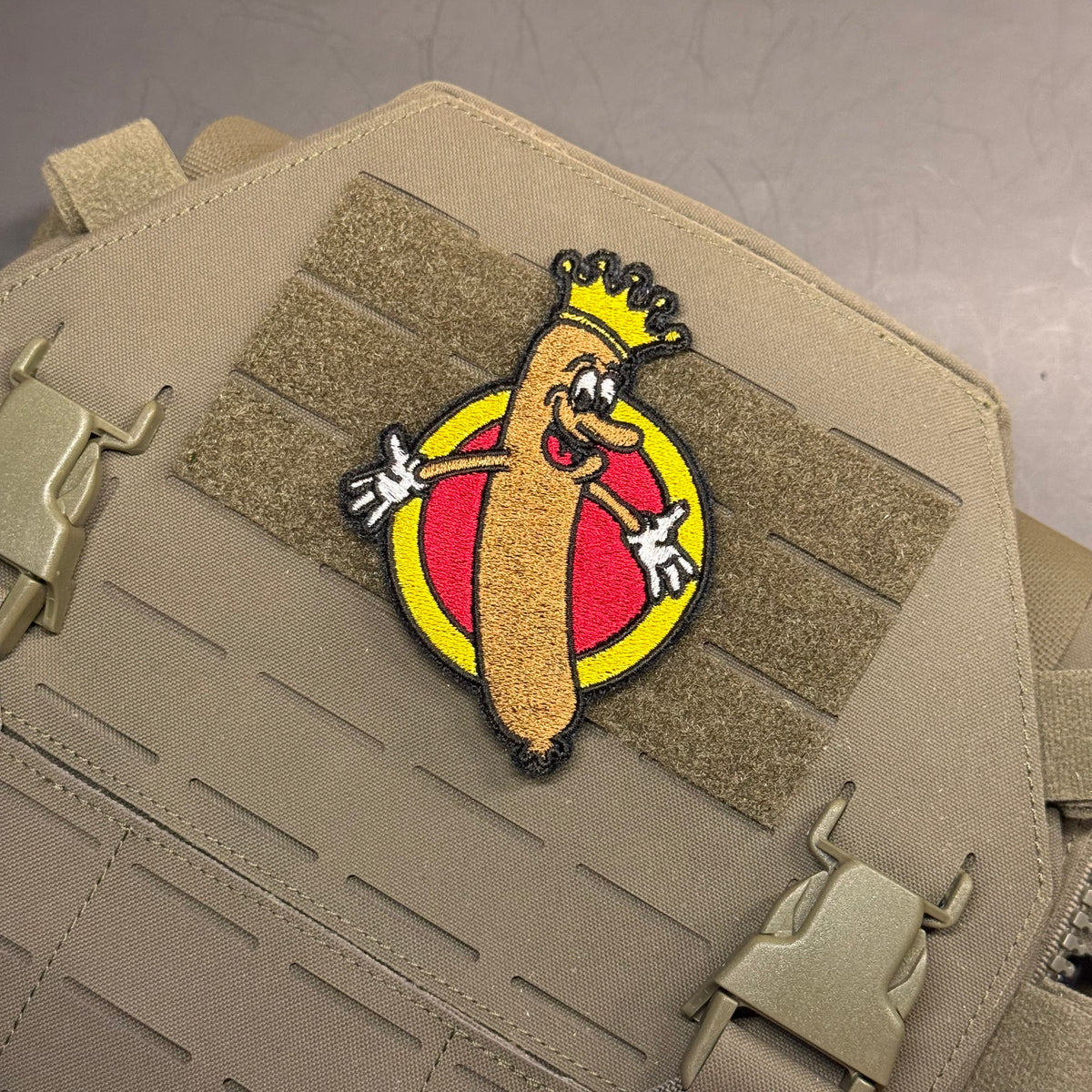 A tactical embroidered patch featuring a cartoon hot dog character wearing a gold crown, set against a red and yellow circular background, attached to a tan tactical vest with MOLLE webbing.