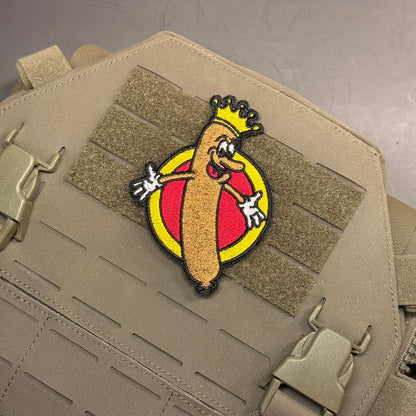 A tactical embroidered patch featuring a cartoon hot dog character wearing a gold crown, set against a red and yellow circular background, attached to a tan tactical vest with MOLLE webbing.