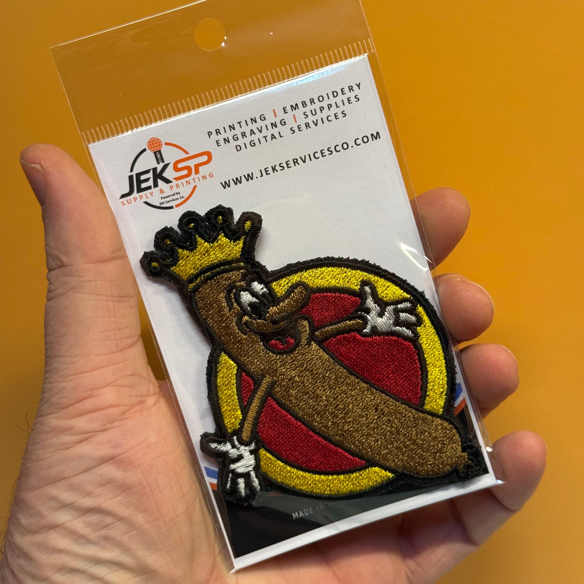 A hand holds a packaged embroidered patch featuring a cartoon banana character wearing a gold crown, with a red and yellow circular background. The patch is mounted on a white backing card printed with the JEKSP logo and text.