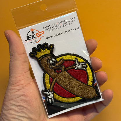 A hand holds a packaged embroidered patch featuring a cartoon banana character wearing a gold crown, with a red and yellow circular background. The patch is mounted on a white backing card printed with the JEKSP logo and text.