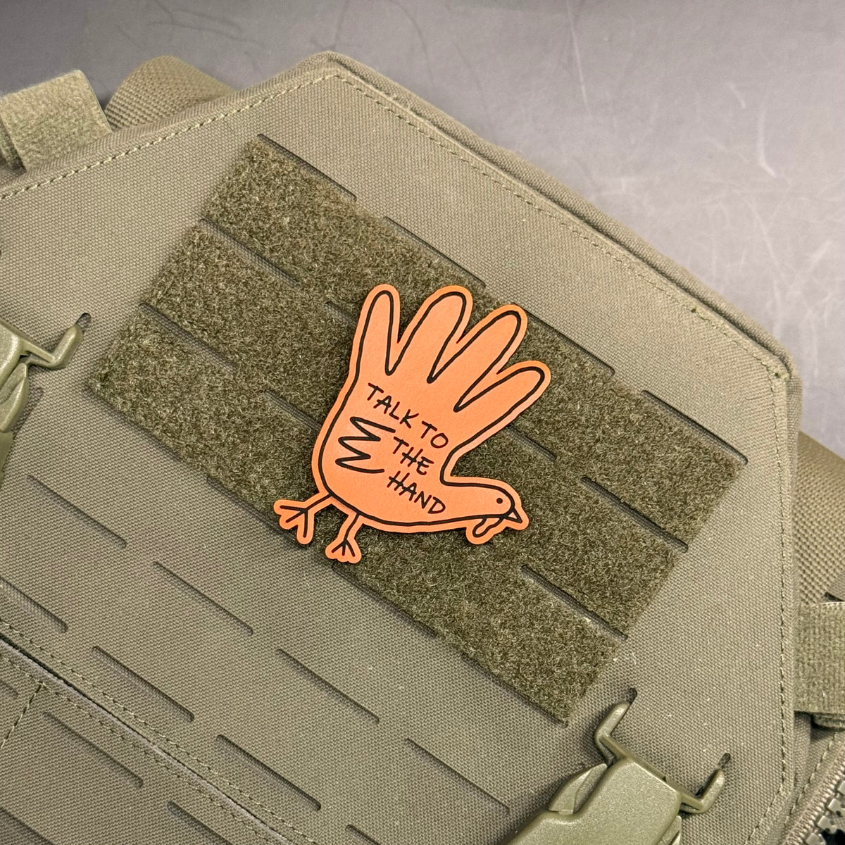 An orange PVC patch shaped like a hand with the text 'TALK TO THE HAND' sewn onto a tactical bag's MOLLE webbing.