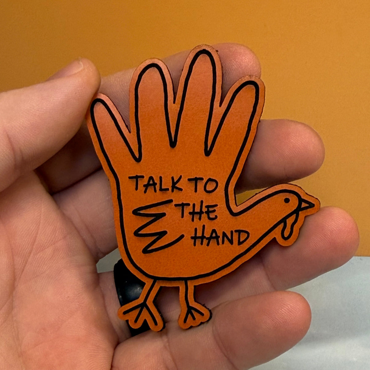 A hand holding a novelty orange hand-shaped ornament or sign shaped like a turkey, with black outline details and the text 'TALK TO THE HAND' on the palm side, against a warm tan background.