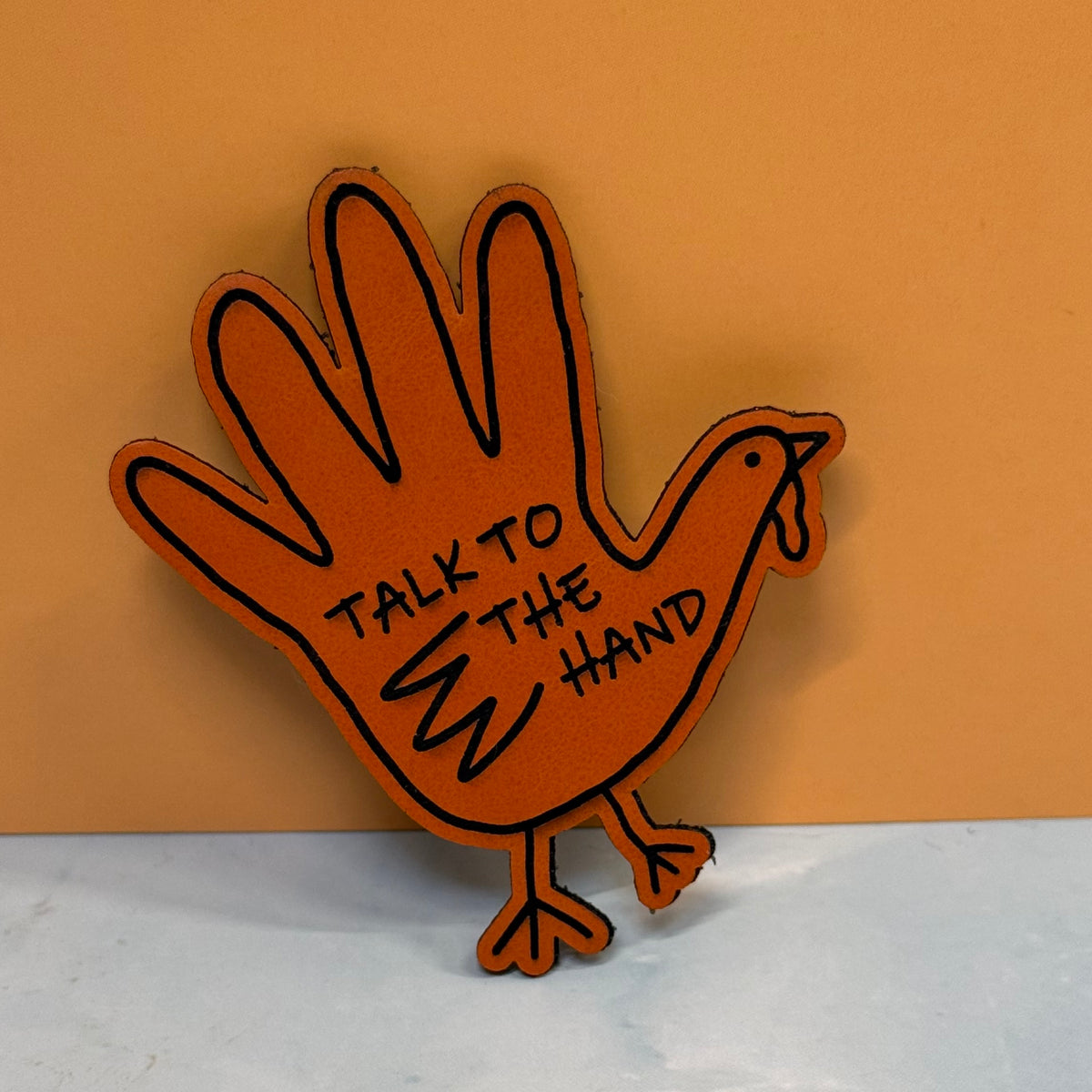 A novelty wooden sign shaped like a turkey hand, painted in a warm orange-brown tone with black outlines and the text 'TALK TO THE HAND' on the palm, set against a solid tan background.