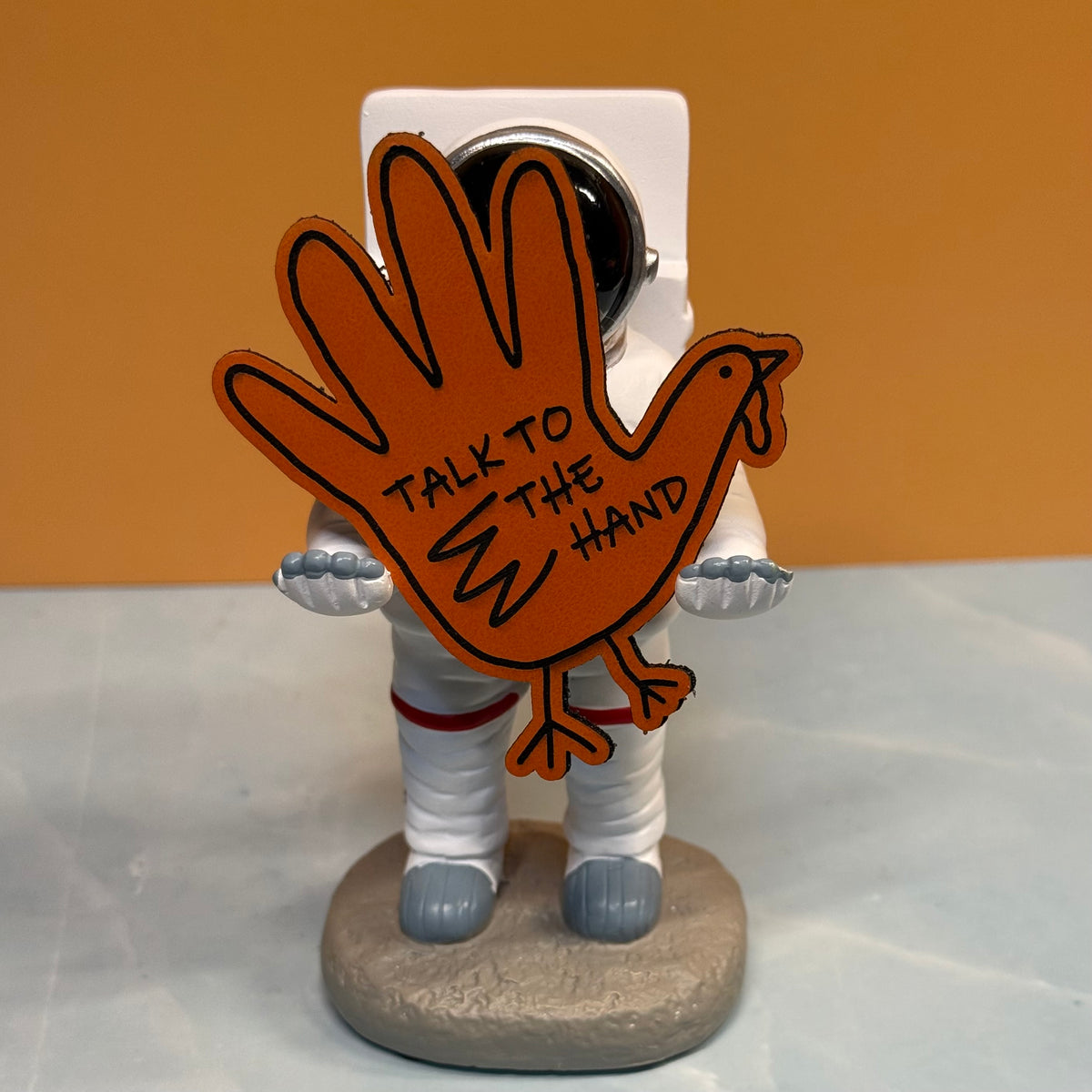 A novelty astronaut figurine holding a large orange hand-shaped sign that reads 'TALK TO THE HAND' in black lettering, set against a warm tan background and light gray surface.