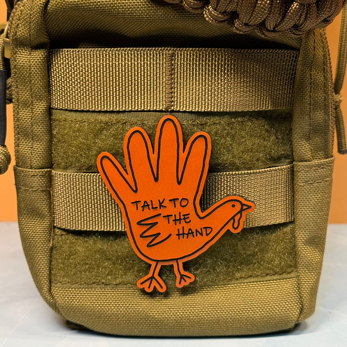An orange rubber patch shaped like a hand with a turkey body and the text 'TALK TO THE HAND' attached to an olive drab tactical pouch.