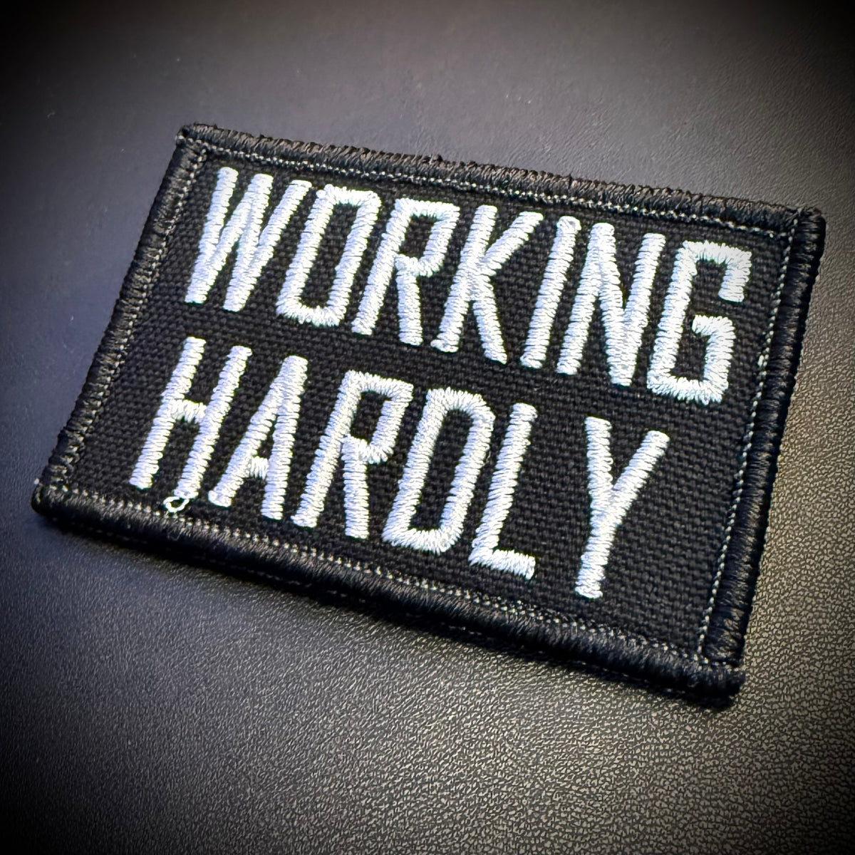A rectangular black embroidered patch with the words 'WORKING HARDLY' in bold white capital letters, shown on a dark textured surface.