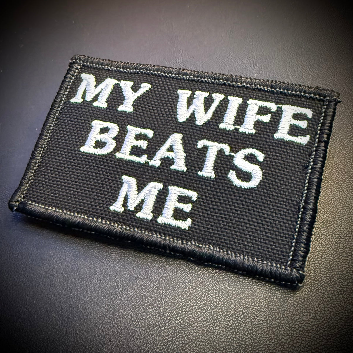 A rectangular black embroidered patch with white text reading 'MY WIFE BEATS ME' on a dark textured background.
