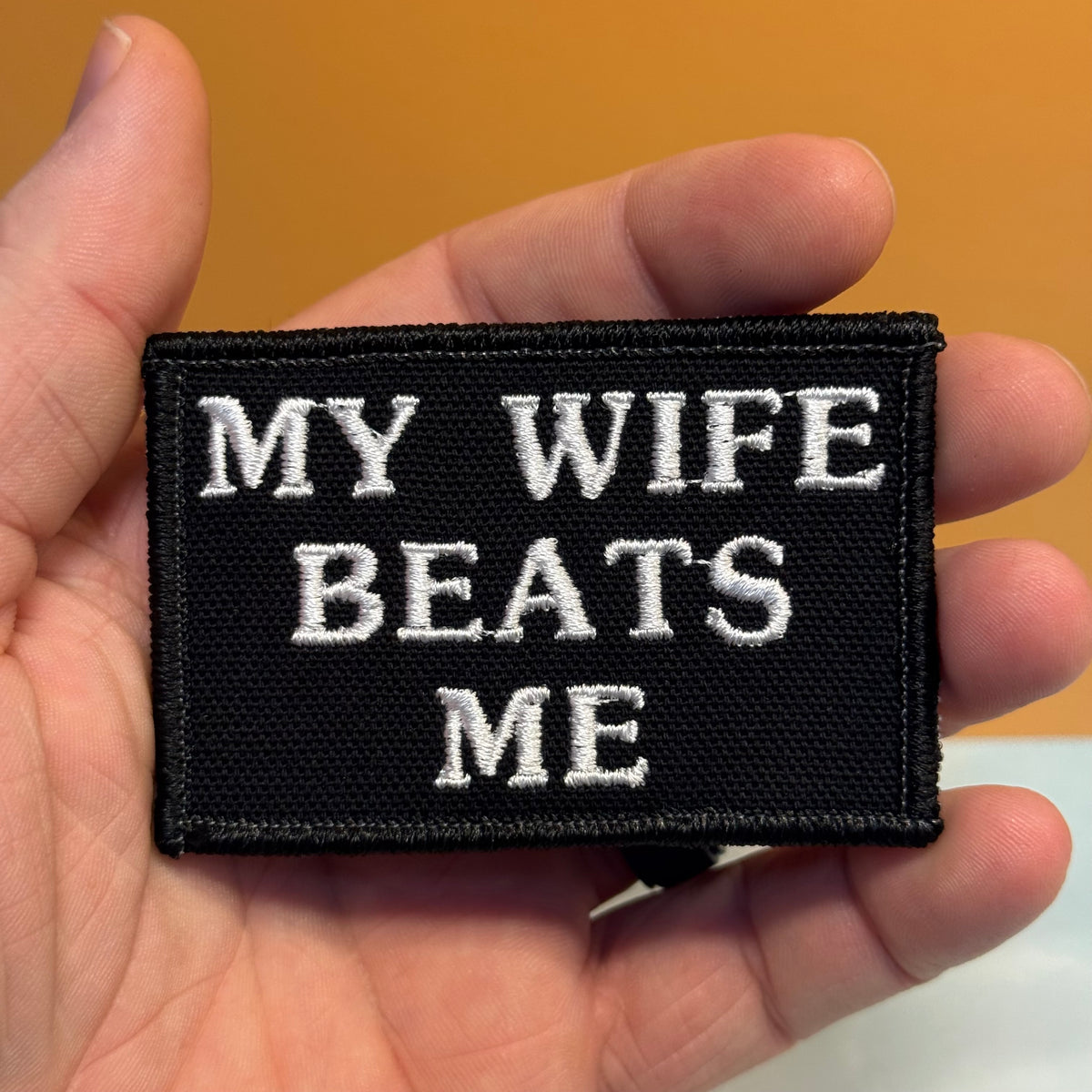 A black rectangular embroidered patch held in a hand, featuring the white text 'MY WIFE BEATS ME' on a warm tan background.