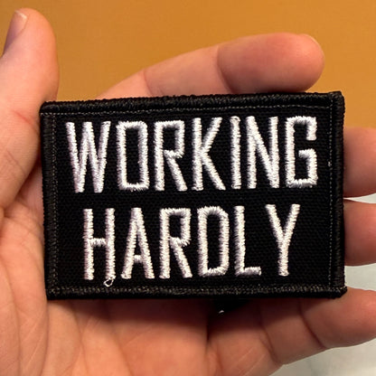 A rectangular black embroidered patch with white text reading 'WORKING HARDLY' is held in a person's hand against a plain tan background.