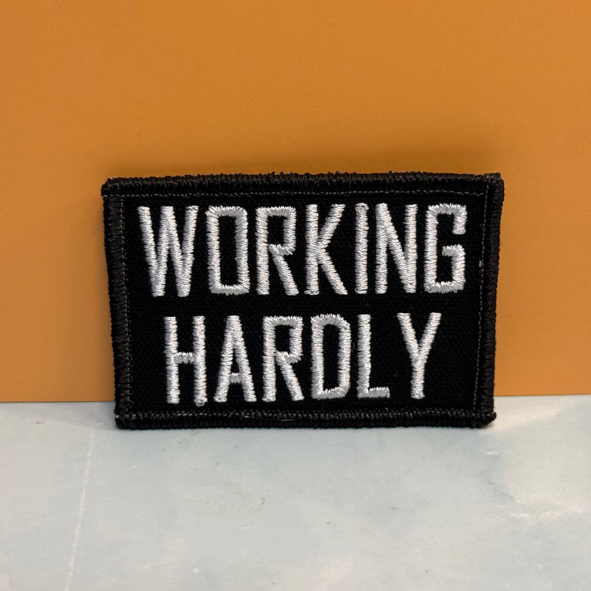 A rectangular black embroidered patch with the words 'WORKING HARDLY' in white capital letters, shown against a warm orange background and light gray surface.