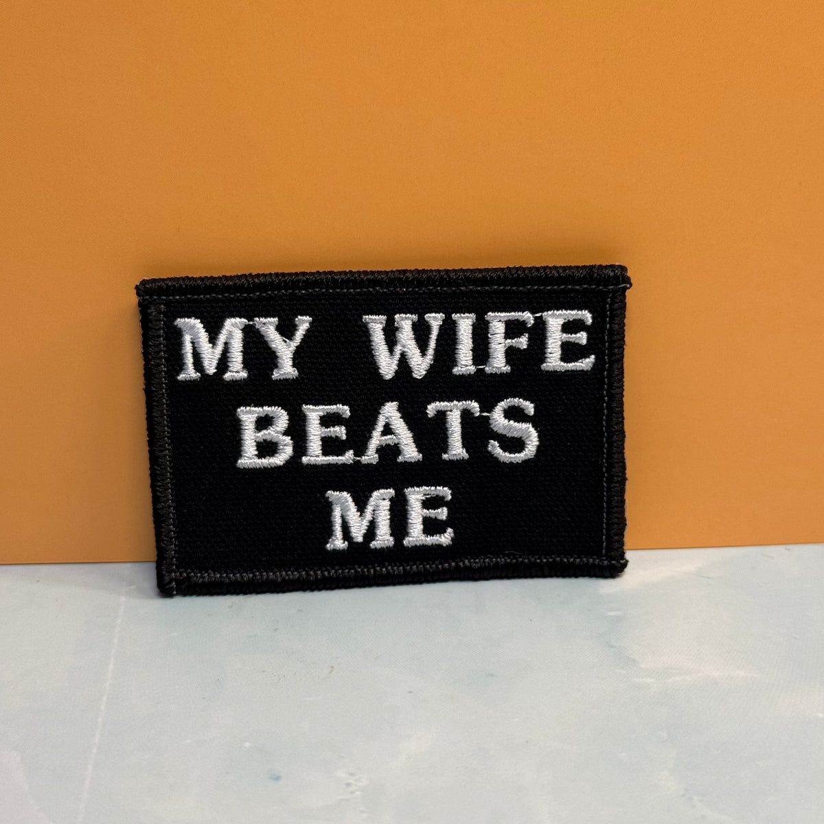 A rectangular black embroidered patch with white text reading 'MY WIFE BEATS ME' against a warm orange background and light gray surface.