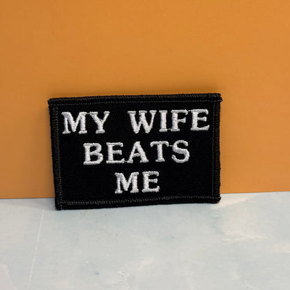 A rectangular black embroidered patch with white text reading 'MY WIFE BEATS ME' against a warm orange background and light gray surface.