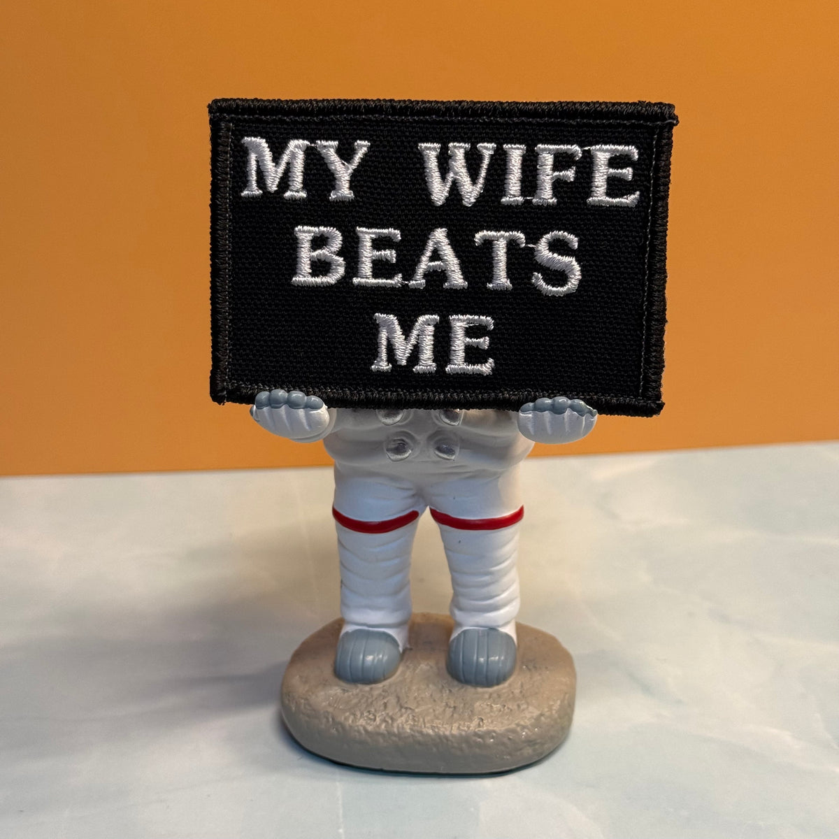A small astronaut figurine holding a black rectangular sign with white embroidered text that reads “MY WIFE BEATS ME,” set against a warm tan background and light surface.