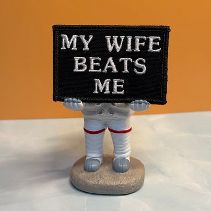 A small astronaut figurine holding a black rectangular sign with white embroidered text that reads “MY WIFE BEATS ME,” set against a warm tan background and light surface.