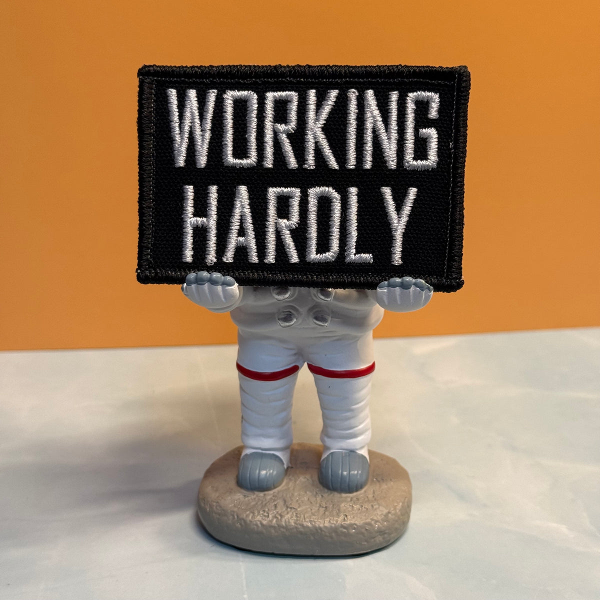 A small astronaut figurine holding a rectangular black patch with white embroidered text reading “WORKING HARDLY,” standing on a round gray base against an orange background.