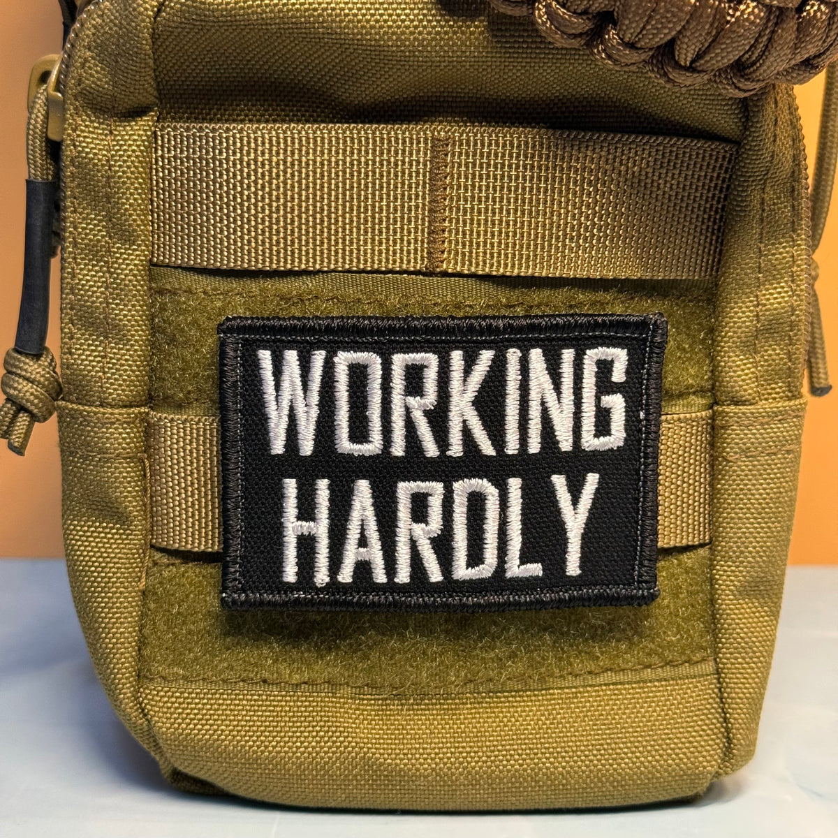 A close-up of a tactical pouch in olive drab green nylon with a black rectangular patch on the front that reads 'WORKING HARDLY' in white capital letters.