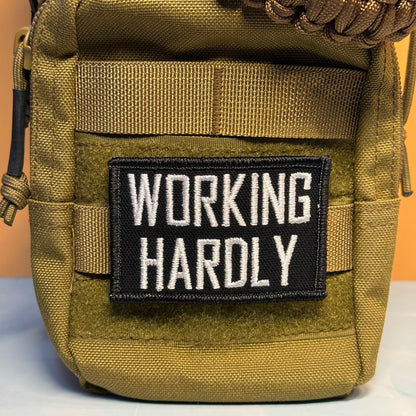 A close-up of a tactical pouch in olive drab green nylon with a black rectangular patch on the front that reads 'WORKING HARDLY' in white capital letters.