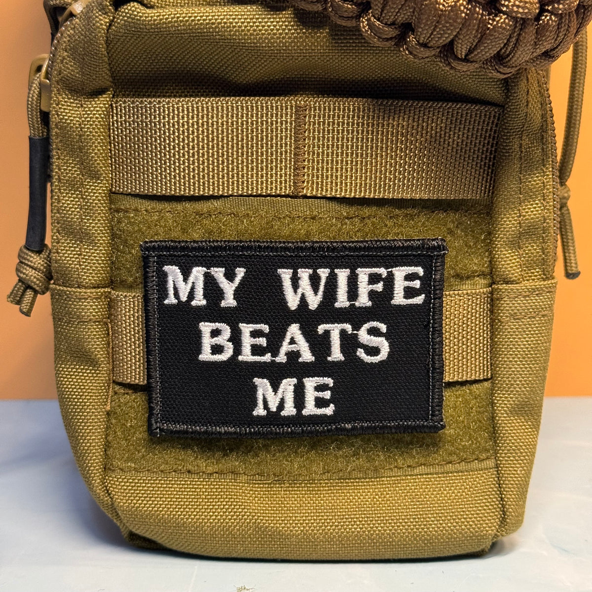 An olive drab tactical pouch with a black rectangular patch featuring white embroidered text that reads 'MY WIFE BEATS ME'. The pouch has MOLLE webbing and a zipper on the left side.
