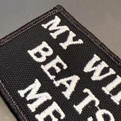 A close-up of a black embroidered patch with white text reading 'MY WILDERNESS BEATS ME' on a dark gray background.