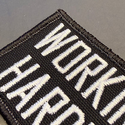 Close-up of a black embroidered patch with bold white text reading 'WORK HARD' on a dark gray background.