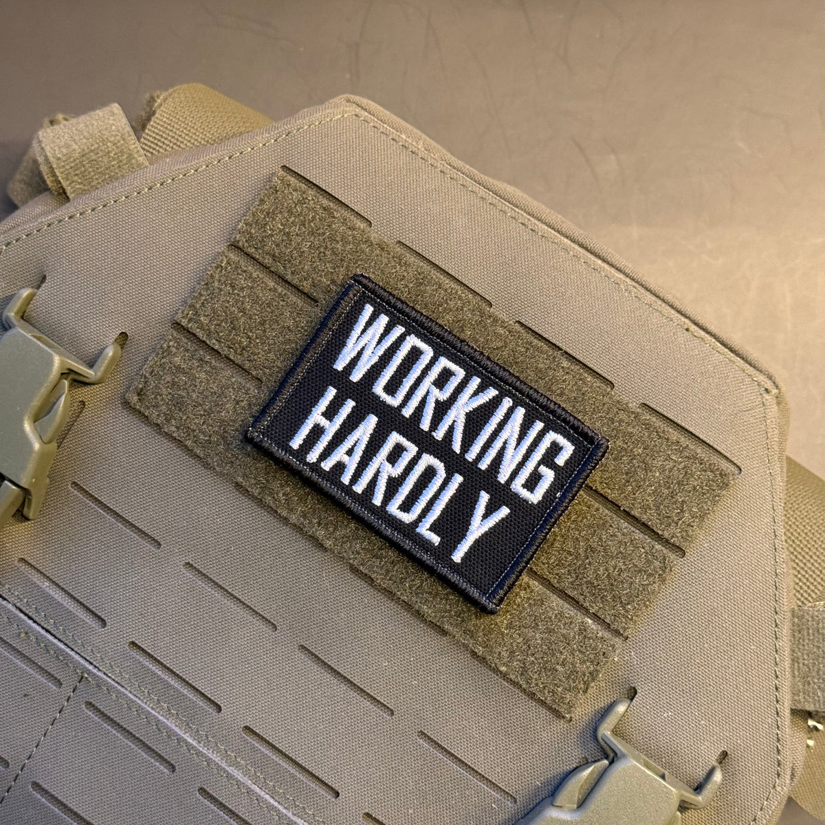 A rectangular black embroidered patch with the words 'WORKING HARDLY' in white capital letters, attached to a tan tactical vest with hook-and-loop fastening.