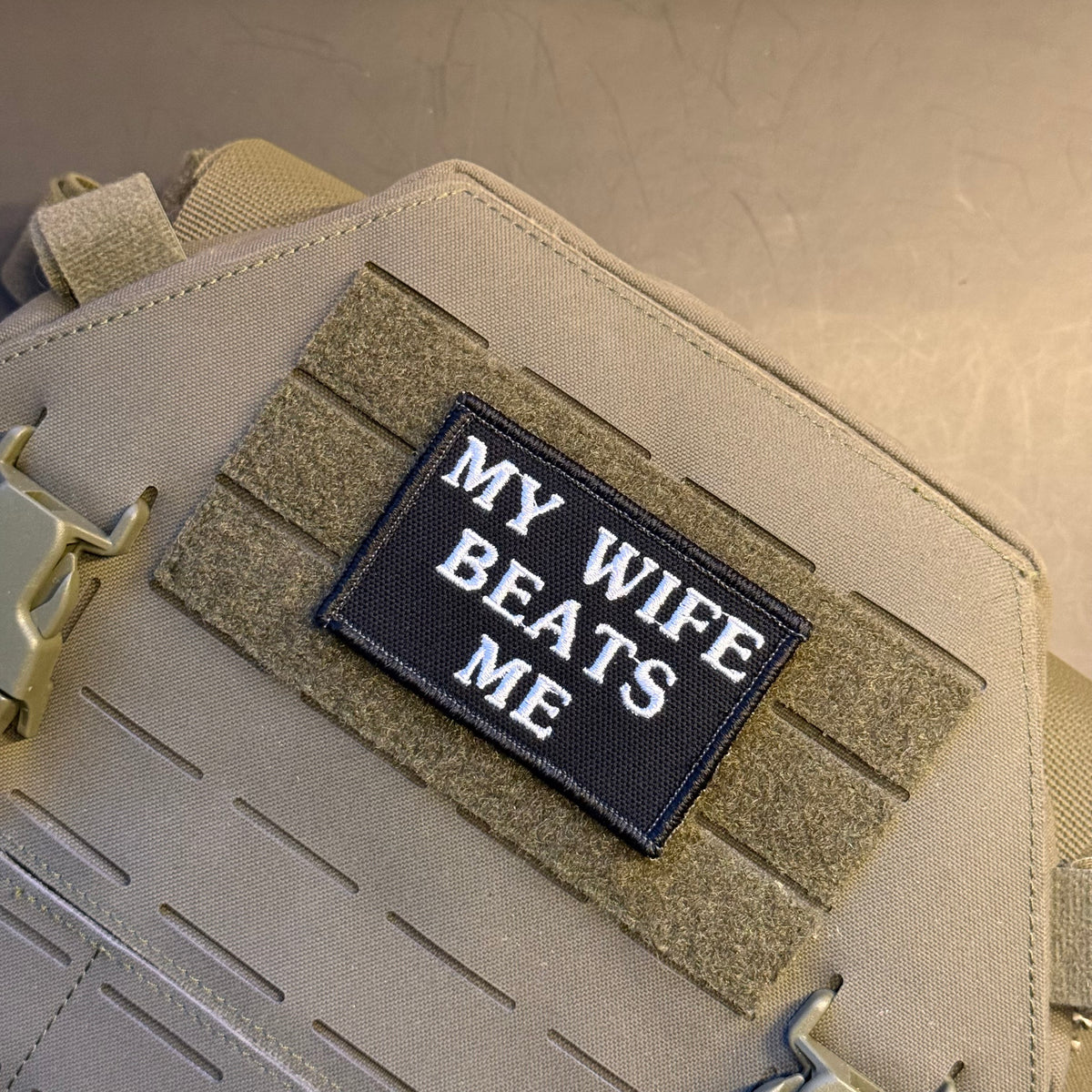 A close-up of a tactical bag or vest in tan canvas with a black rectangular embroidered patch reading 'MY WIFE BEATS ME' in white letters, attached to tan Velcro-style strips.