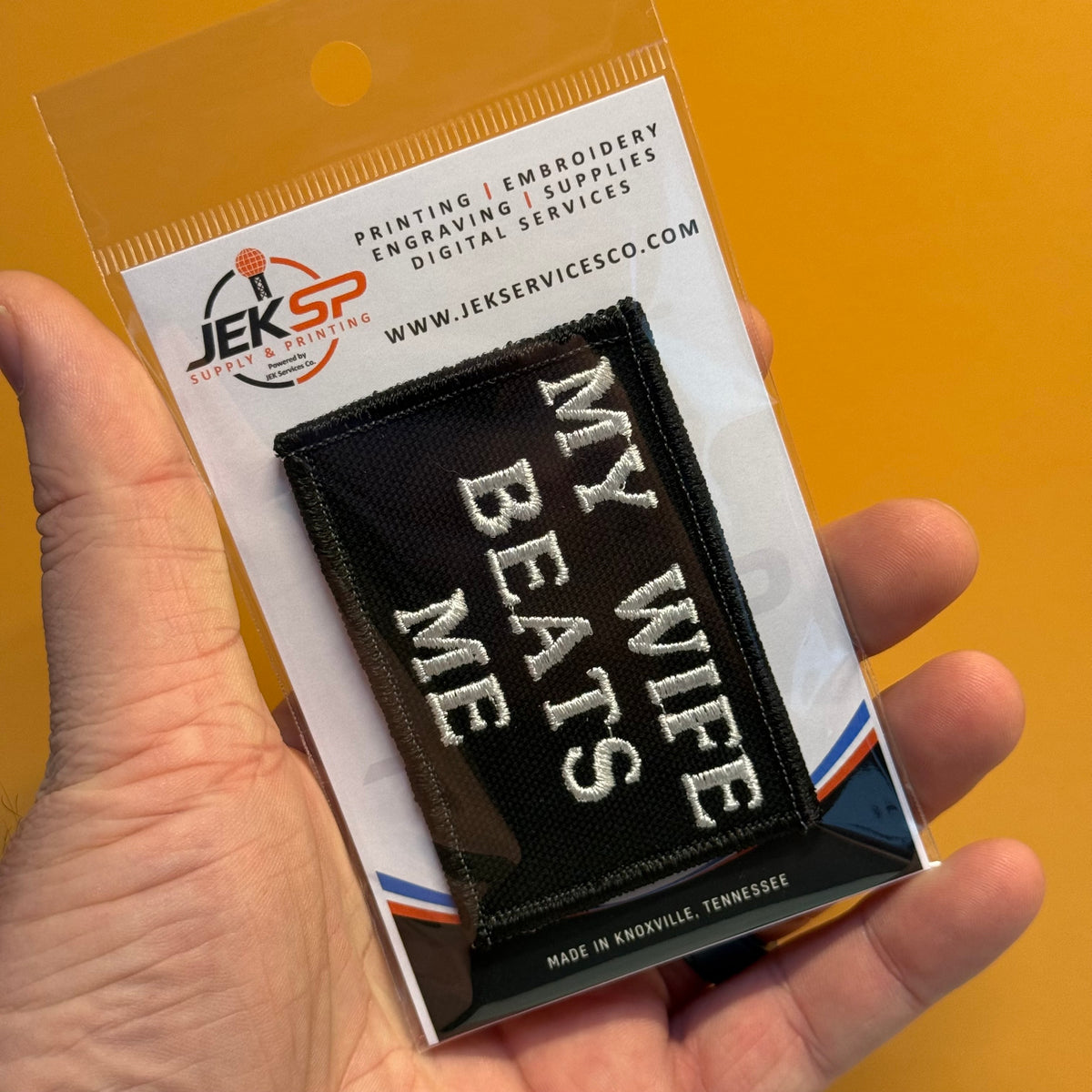 A hand holds a packaged black embroidered patch that reads “MY WIFE BEATS ME” in white lettering, mounted on a branded Jeksp Service & Supply card against a warm tan background.