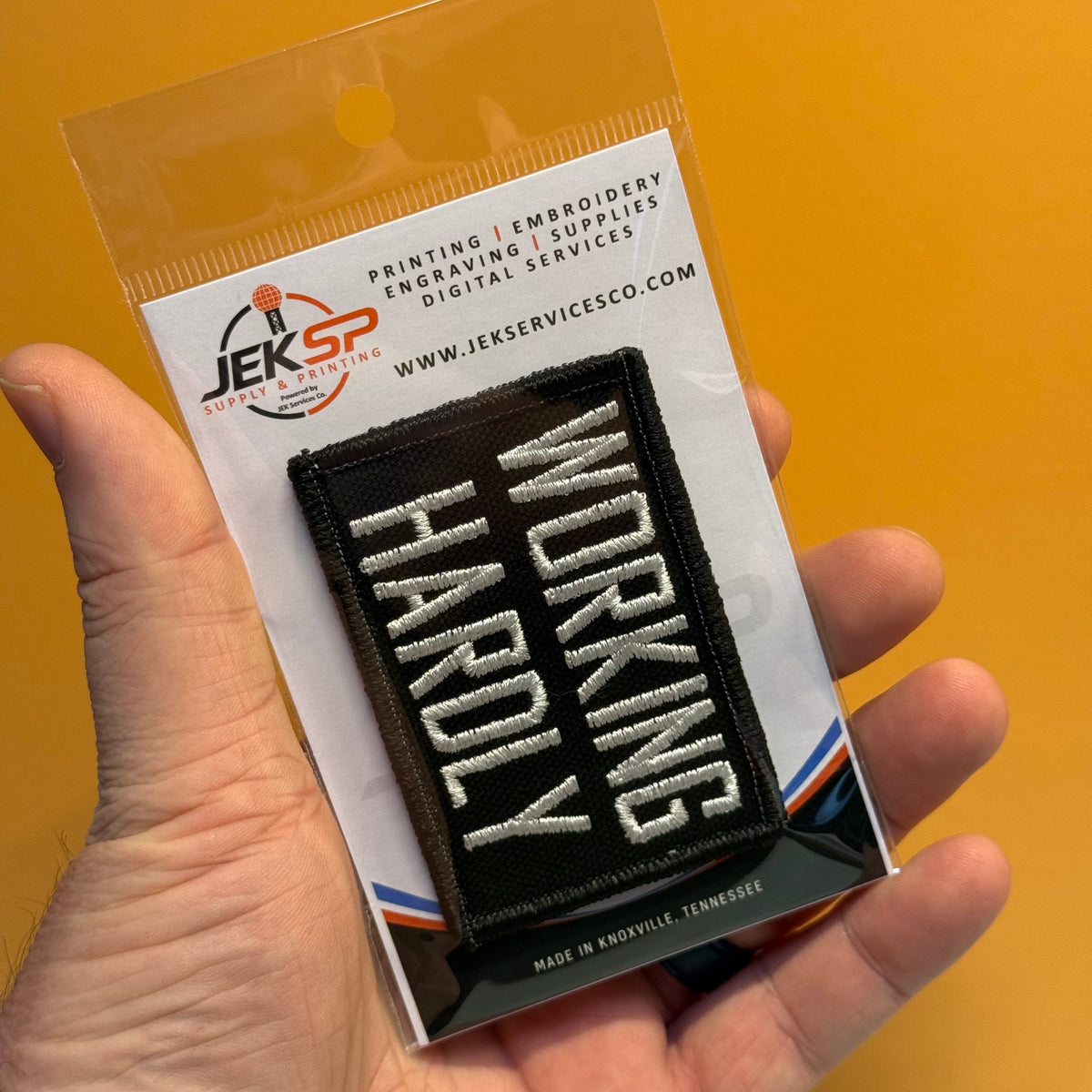 A hand holds a packaged embroidered patch that reads “WORKING HARDLY” in white letters on a black background. The product is shown in clear retail packaging with a printed backing card, against a solid orange background.