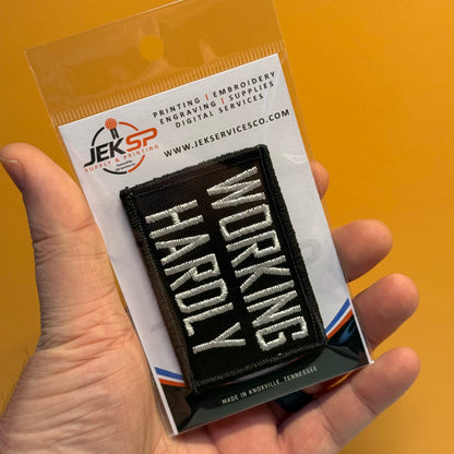 A hand holds a packaged embroidered patch that reads “WORKING HARDLY” in white letters on a black background. The product is shown in clear retail packaging with a printed backing card, against a solid orange background.