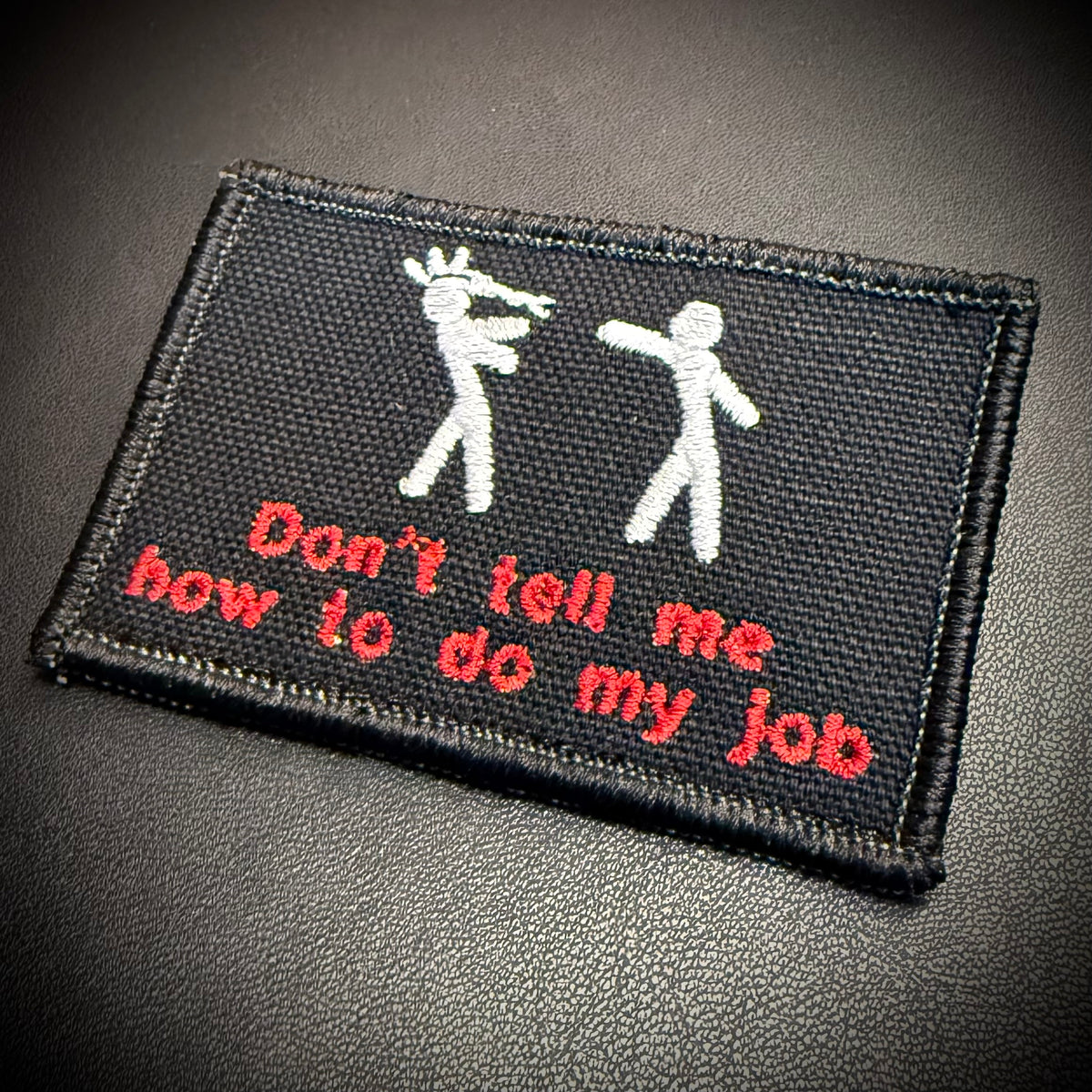 A rectangular black embroidered patch with white stick figures and red text reading 'Don't tell me how to do my job', shown on a dark textured background.