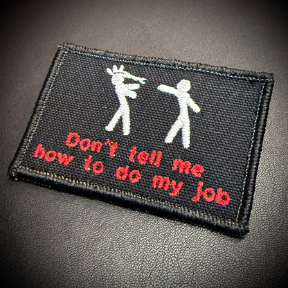 A rectangular black embroidered patch with white stick figures and red text reading 'Don't tell me how to do my job', shown on a dark textured background.