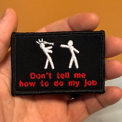 A hand holding a rectangular black embroidered patch with two white stick figures and red text that reads, 'Don't tell me how to do my job.'.