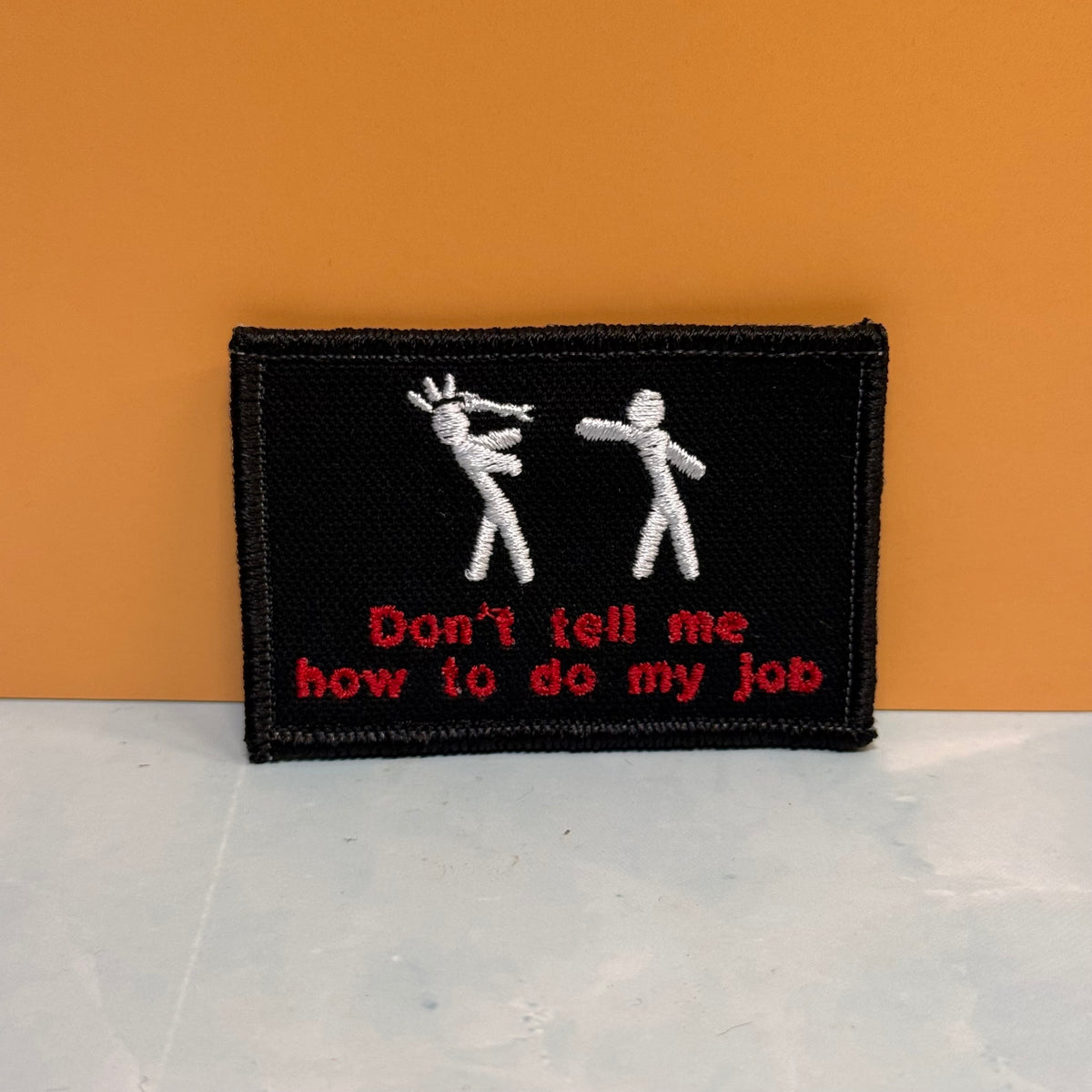 A rectangular black embroidered patch with two white stick figures and red text reading 'Don't tell me how to do my job', shown against an orange wall and light gray surface.