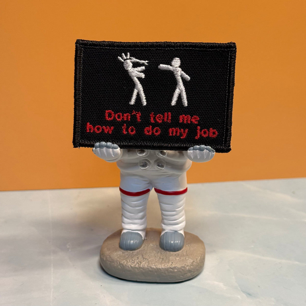 A small figurine of an astronaut holding a black embroidered patch with white stick figures and red text reading 'Don't tell me how to do my job,' set against an orange background and light gray surface.